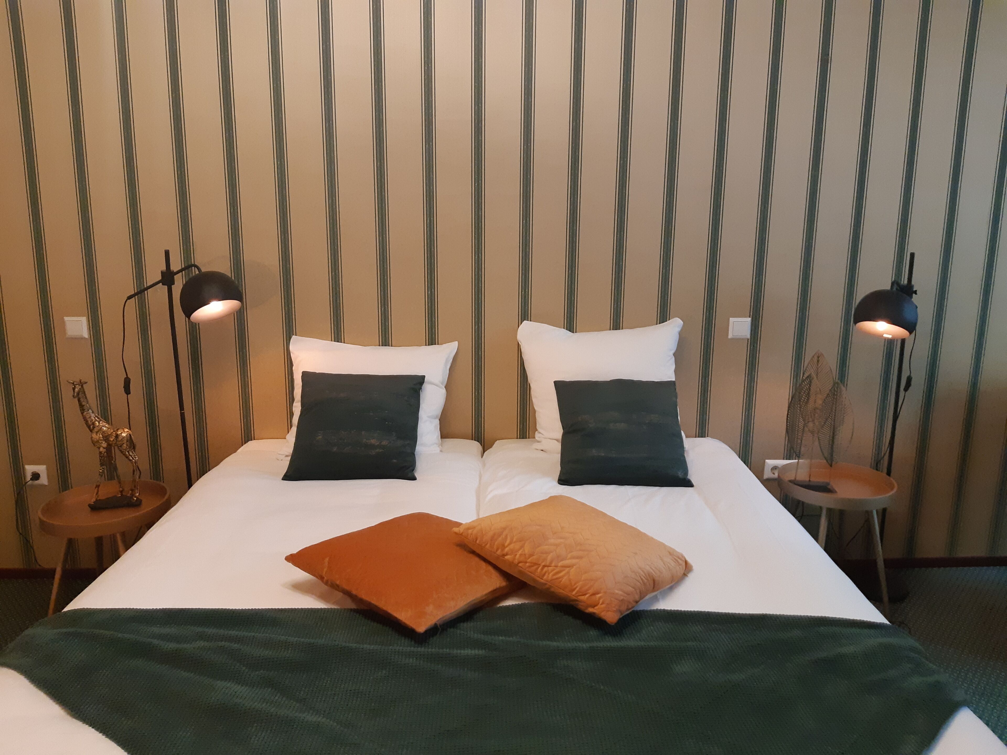 A neatly made bed with white linens and two black pillows, a green throw blanket, and a brown pillow. There are two bedside lamps and a small table with a decorative item on it.
