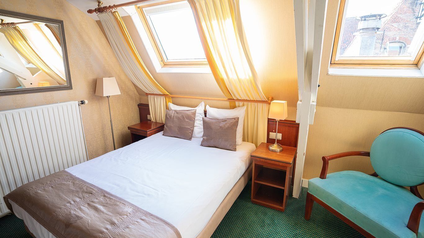 A hotel room with a bed, two bedside tables, a chair, a window with curtains, and a radiator.