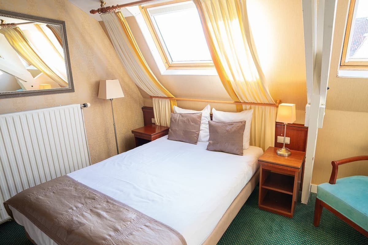 Habitación de hotel con cama, dos mesitas de noche, una silla, ventana con cortinas y un radiador.