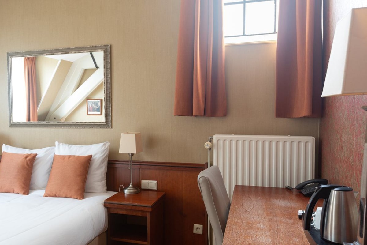 Hotelkamer met een bed, nachtkastjes, een spiegel, een radiator, een tafel met een waterkoker, en gordijnen.