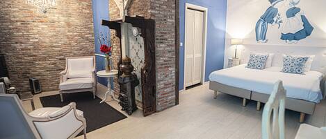 A bedroom with a bed, a chair, a small table, a wall-mounted heater, and a decorative wall piece.