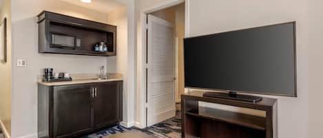 A modern hotel room with a kitchenette, a flat-screen TV, and a patterned carpet.