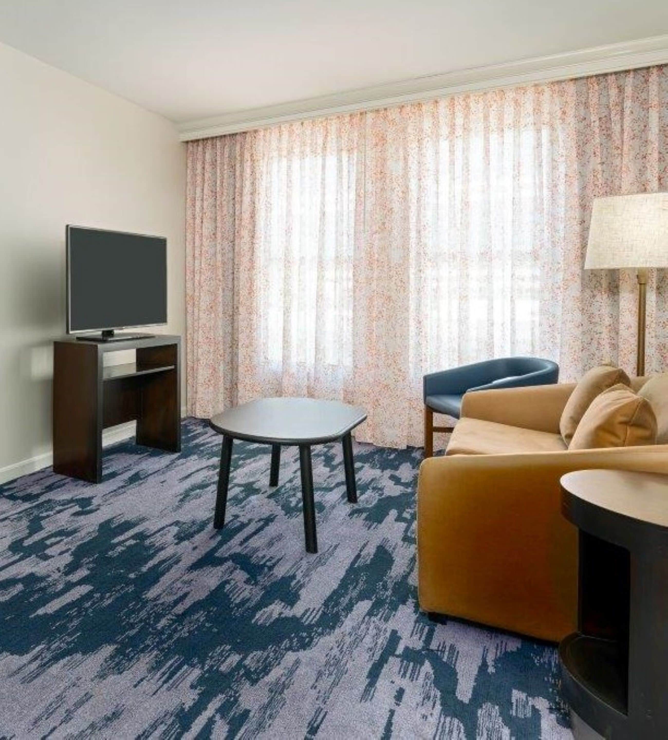 Une chambre d’hôtel équipée d’un téléviseur à écran plat posé sur un meuble en bois, d’une table ronde, d’un canapé et d’une lampe.