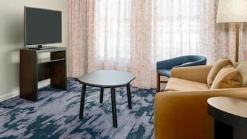 Une chambre d’hôtel équipée d’un téléviseur à écran plat posé sur un meuble en bois, d’une table ronde, d’un canapé et d’une lampe.
