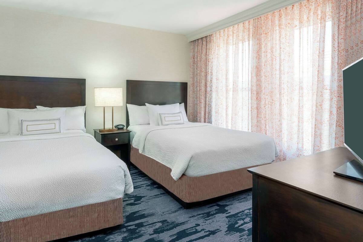 A hotel room with two beds, a desk, a lamp, and a TV.