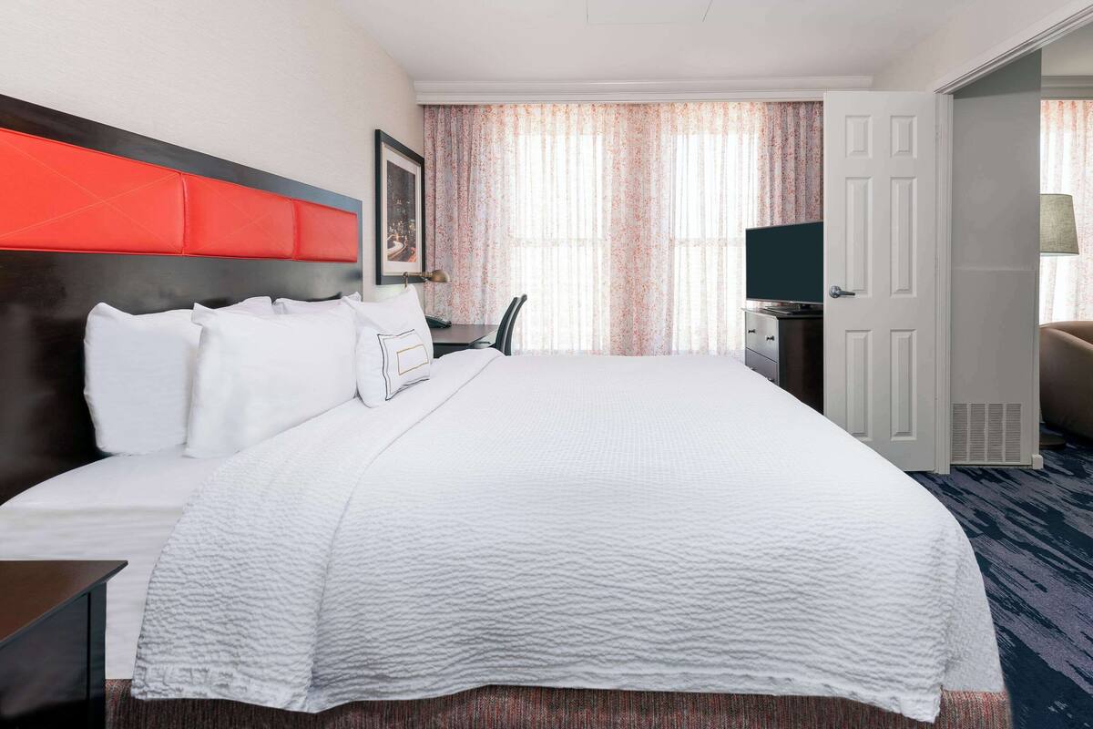 A hotel room with a large bed, a desk with a chair, a television, and a door.