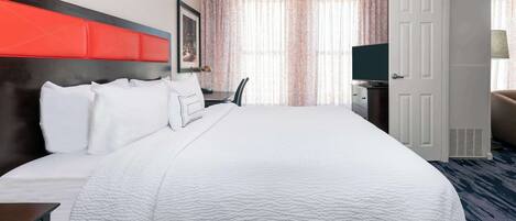 A hotel room with a large bed, a desk with a chair, a television, and a door.