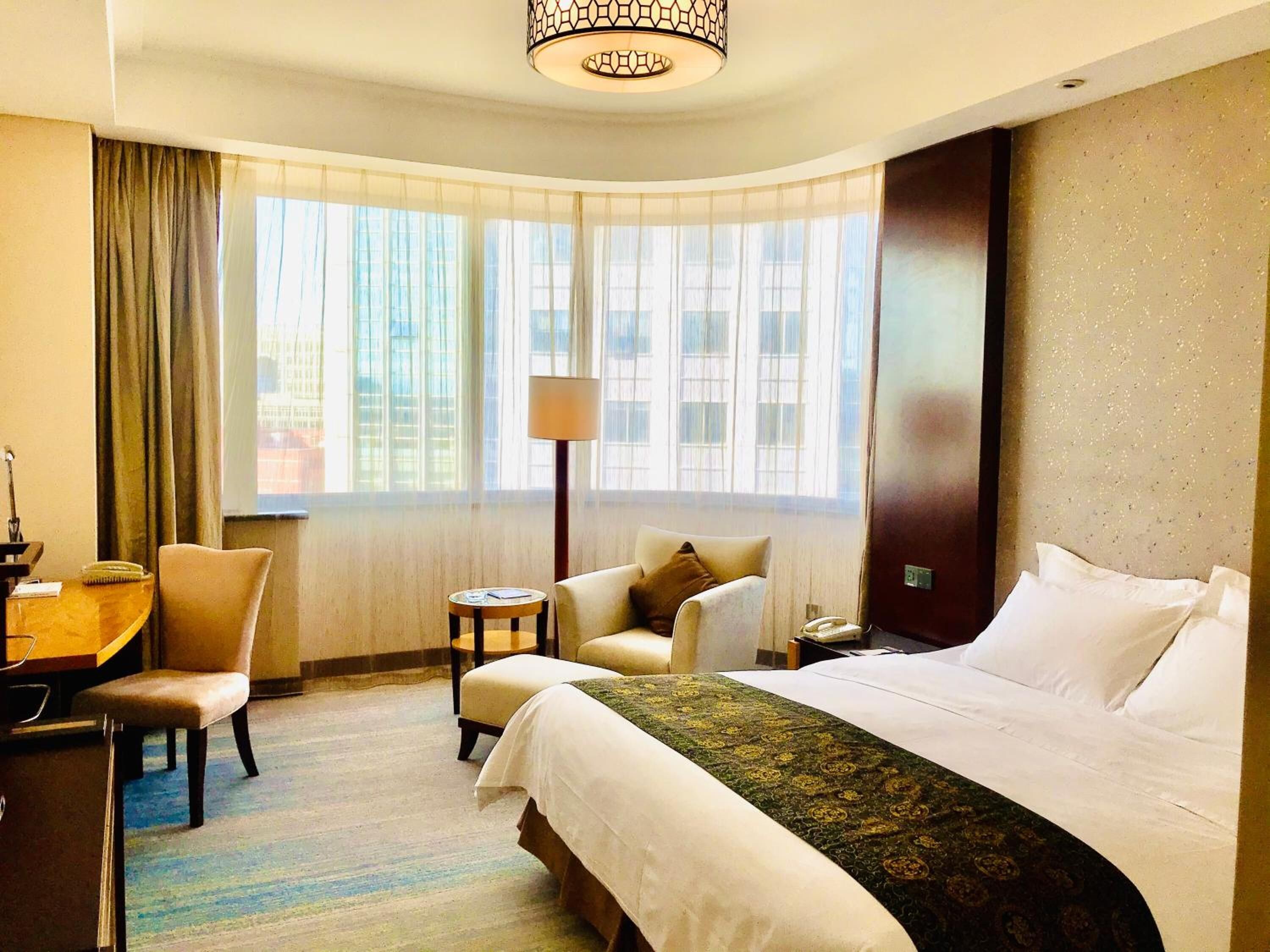 A hotel room with a large bed, a desk, a chair, a lamp, and a view of buildings through the window.