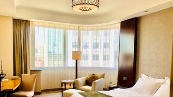 A hotel room with a large bed, a desk, a chair, a lamp, and a view of buildings through the window.