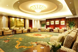Um hall de hotel espaçoso, com um tapete grande e ricamente decorado, áreas de estar confortáveis e um lustre imponente.