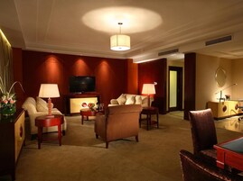 Una habitación de hotel con sofá, sillones, televisor y escritorio con lámpara.