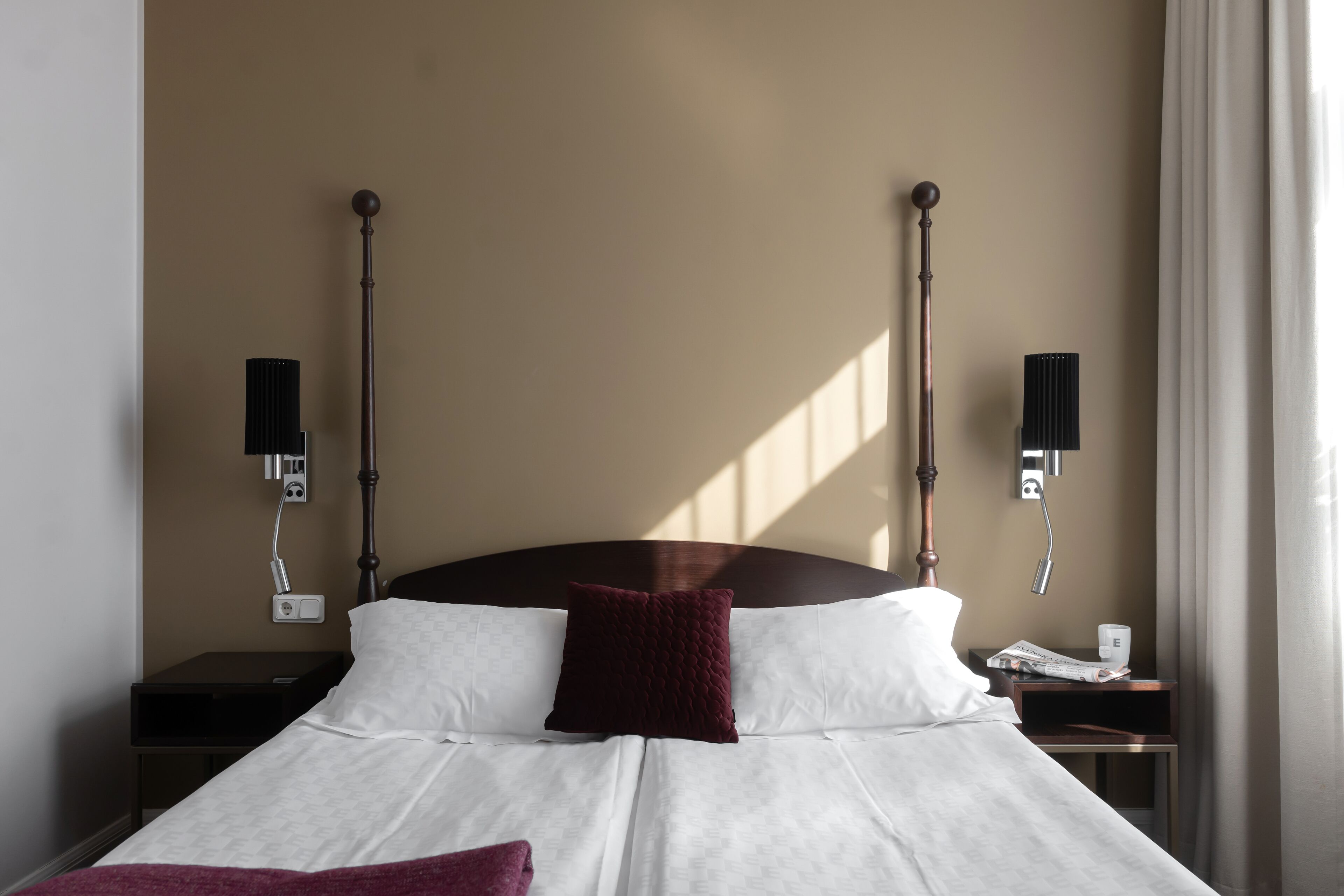 A neatly made bed with white linens and a dark red pillow, flanked by two bedside tables with lamps and a telephone.