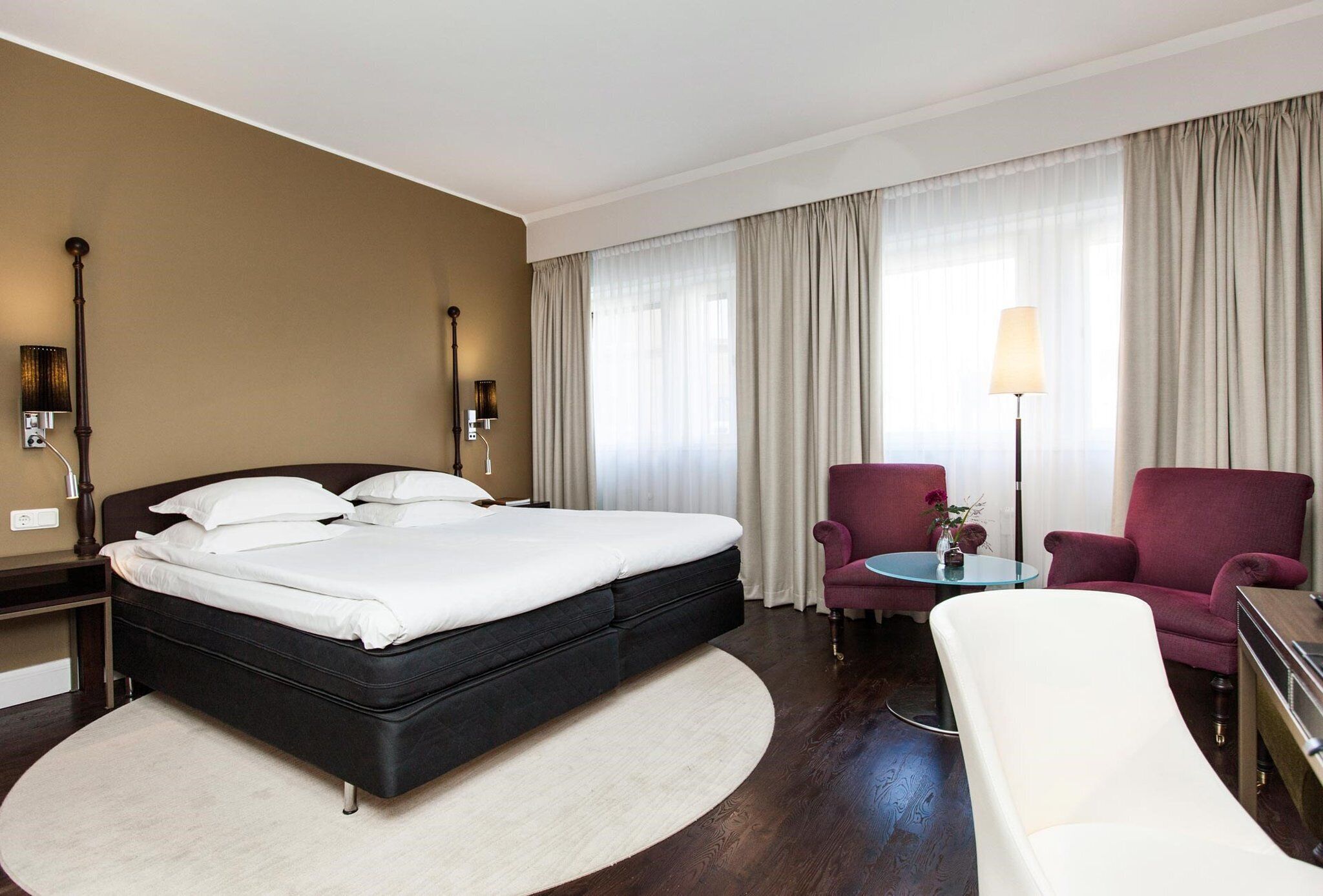 Habitación de hotel con una cama grande, dos sillones, una mesita con un florero y una lámpara.