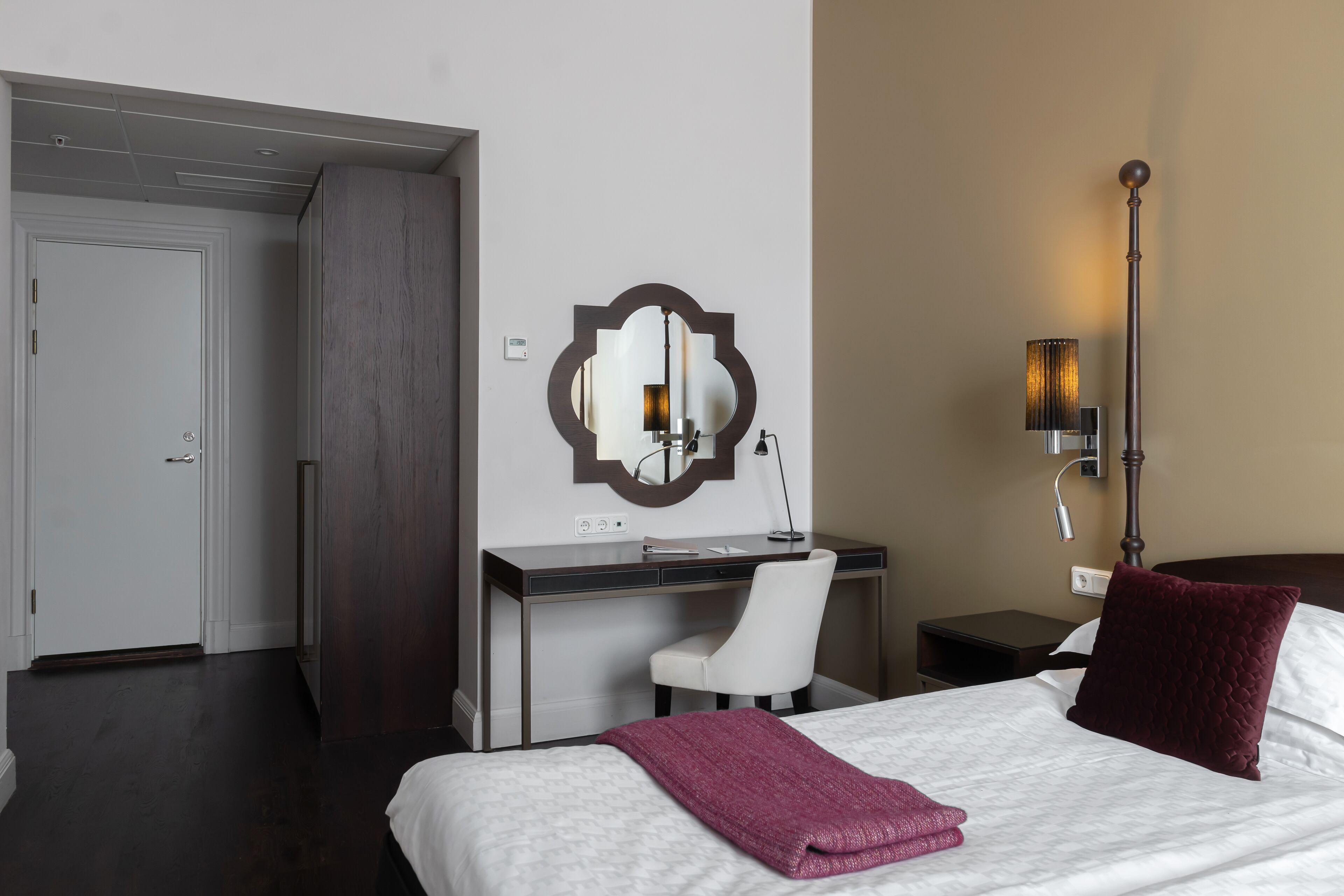 A hotel room with a bed, a desk, a chair, a nightstand, a lamp, and a mirror.