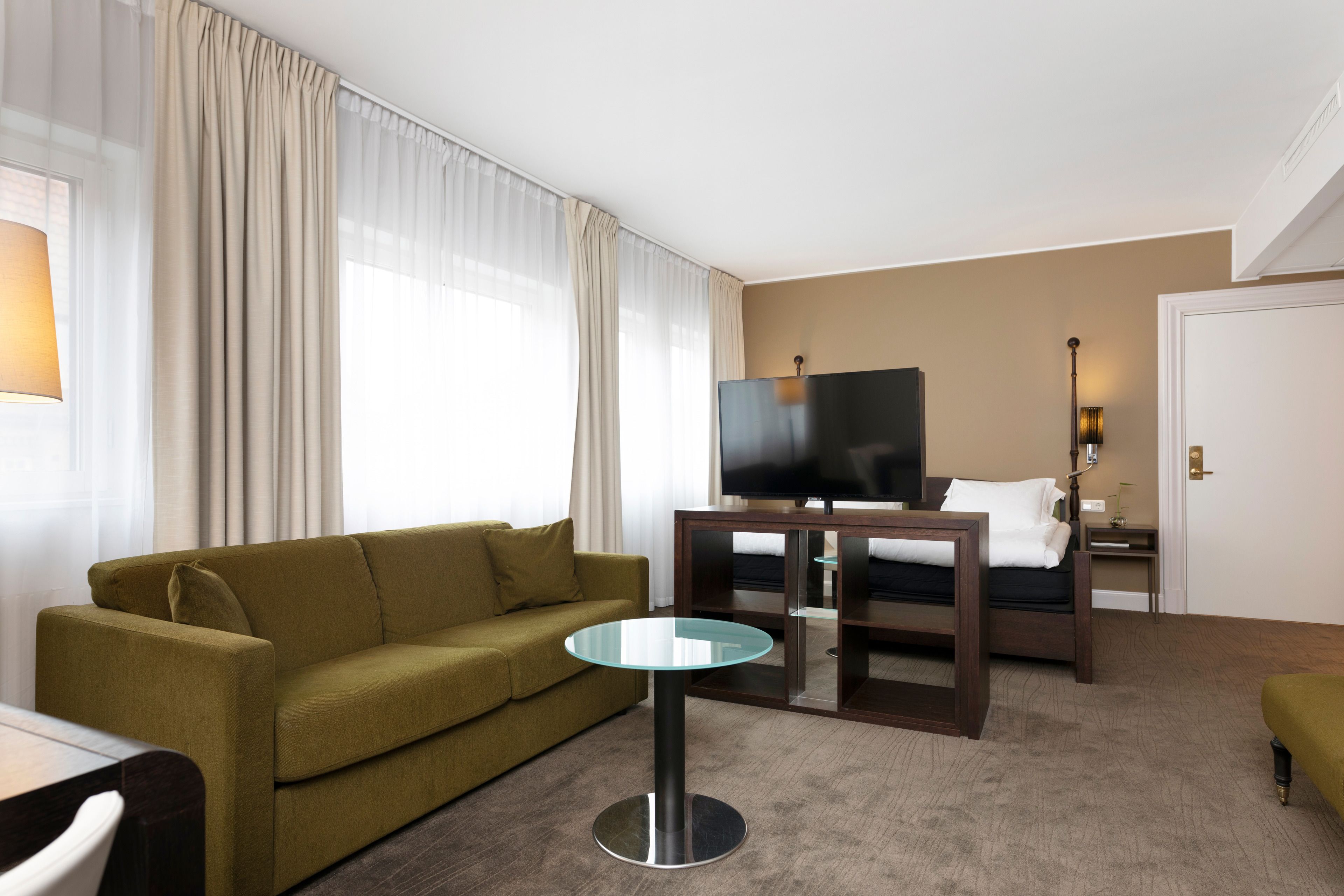 Habitación de hotel con un sofá verde, una mesa de vidrio, una cama con televisor y una mesita de noche con lámpara.