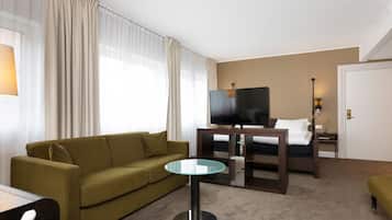 Habitación de hotel con un sofá verde, una mesa de vidrio, una cama con televisor y una mesita de noche con lámpara.