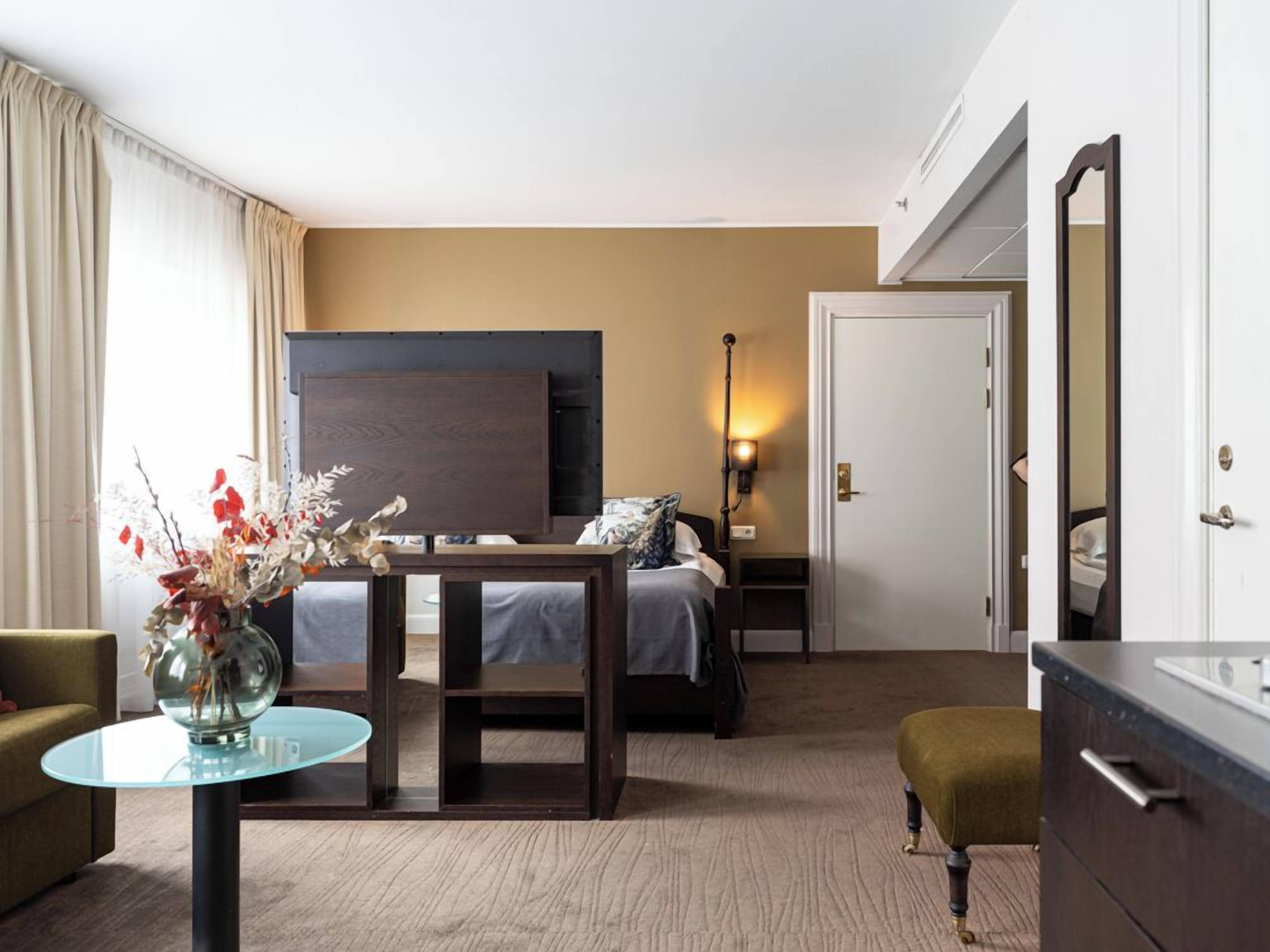 Habitación de hotel con cama, mesita de noche, lámpara, espejo y una pequeña mesa redonda con un florero.
