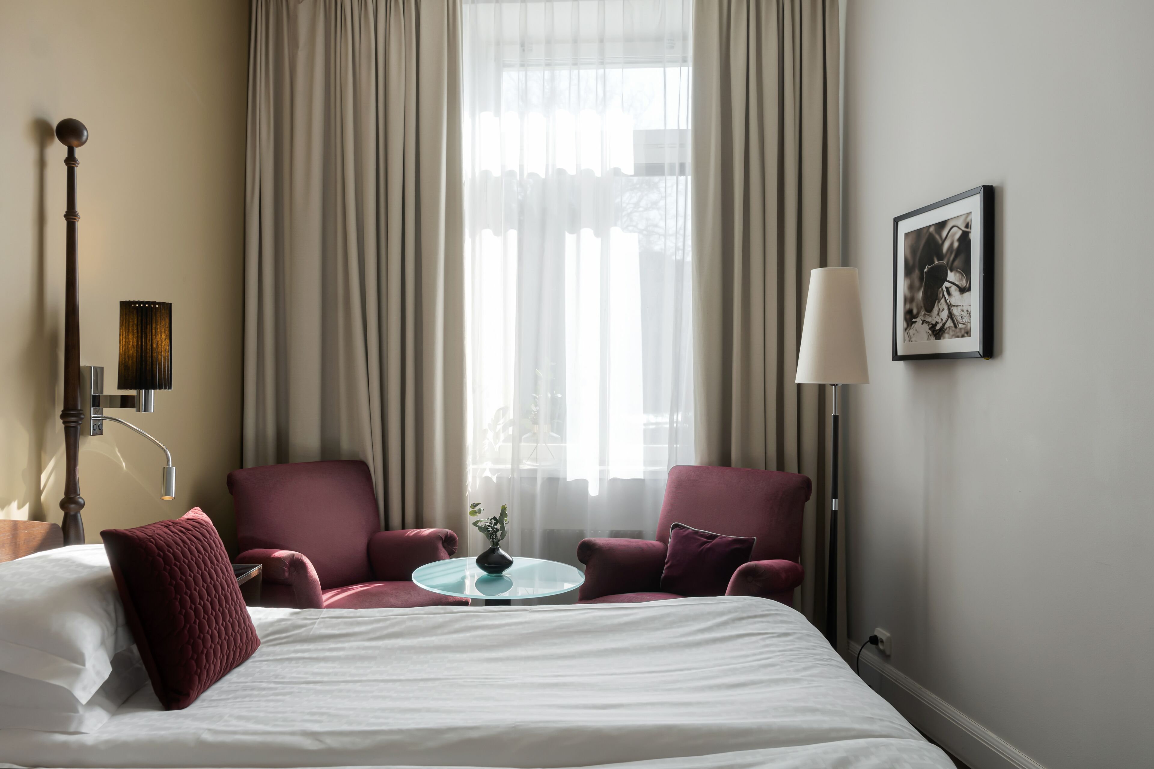 A hotel room with a bed, two armchairs, a small table with a vase, a lamp, and a framed picture on the wall.