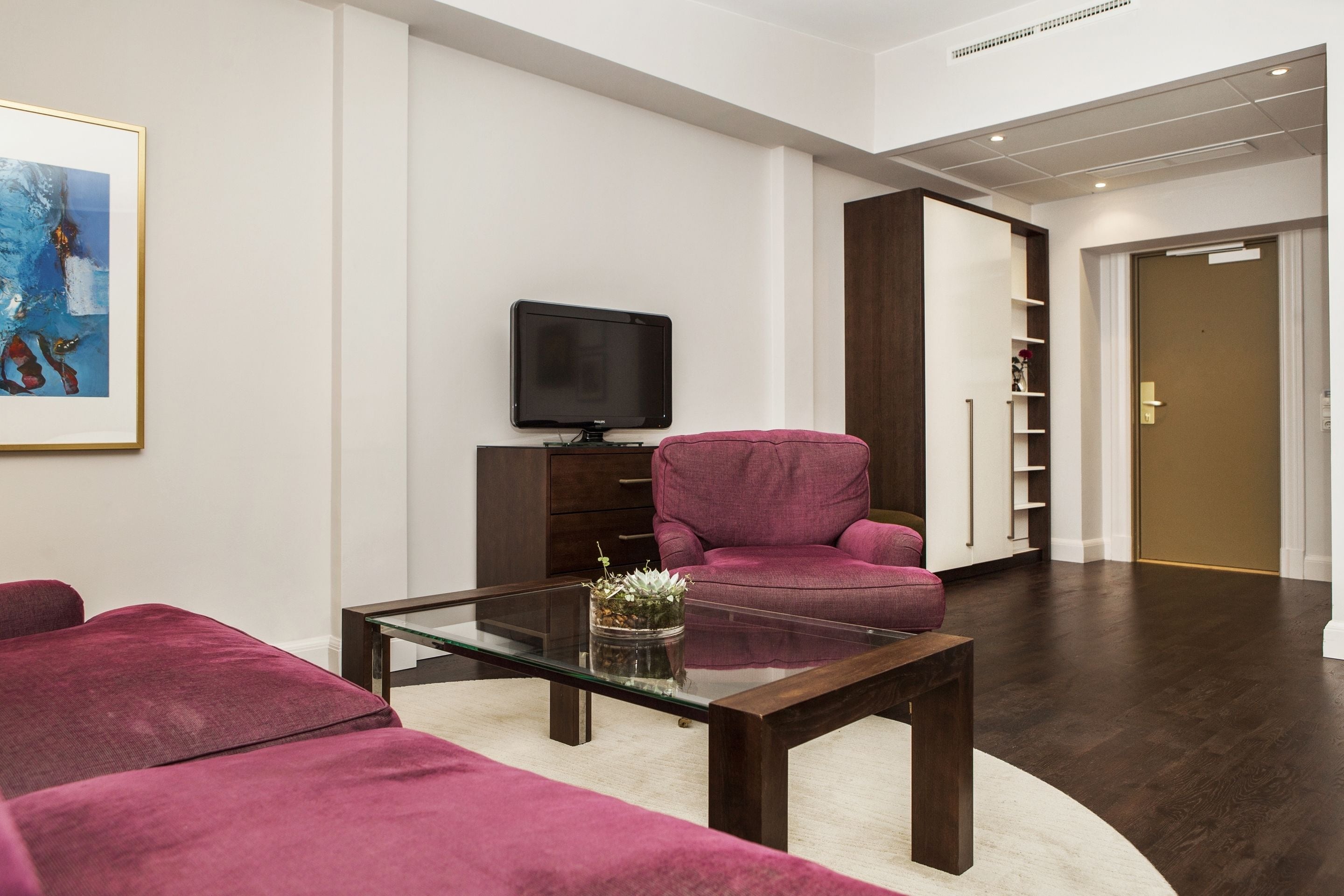 Una sala de estar moderna con una mesa de centro de vidrio, un televisor de pantalla plana, un sillón rosa y un mueble de madera oscura.