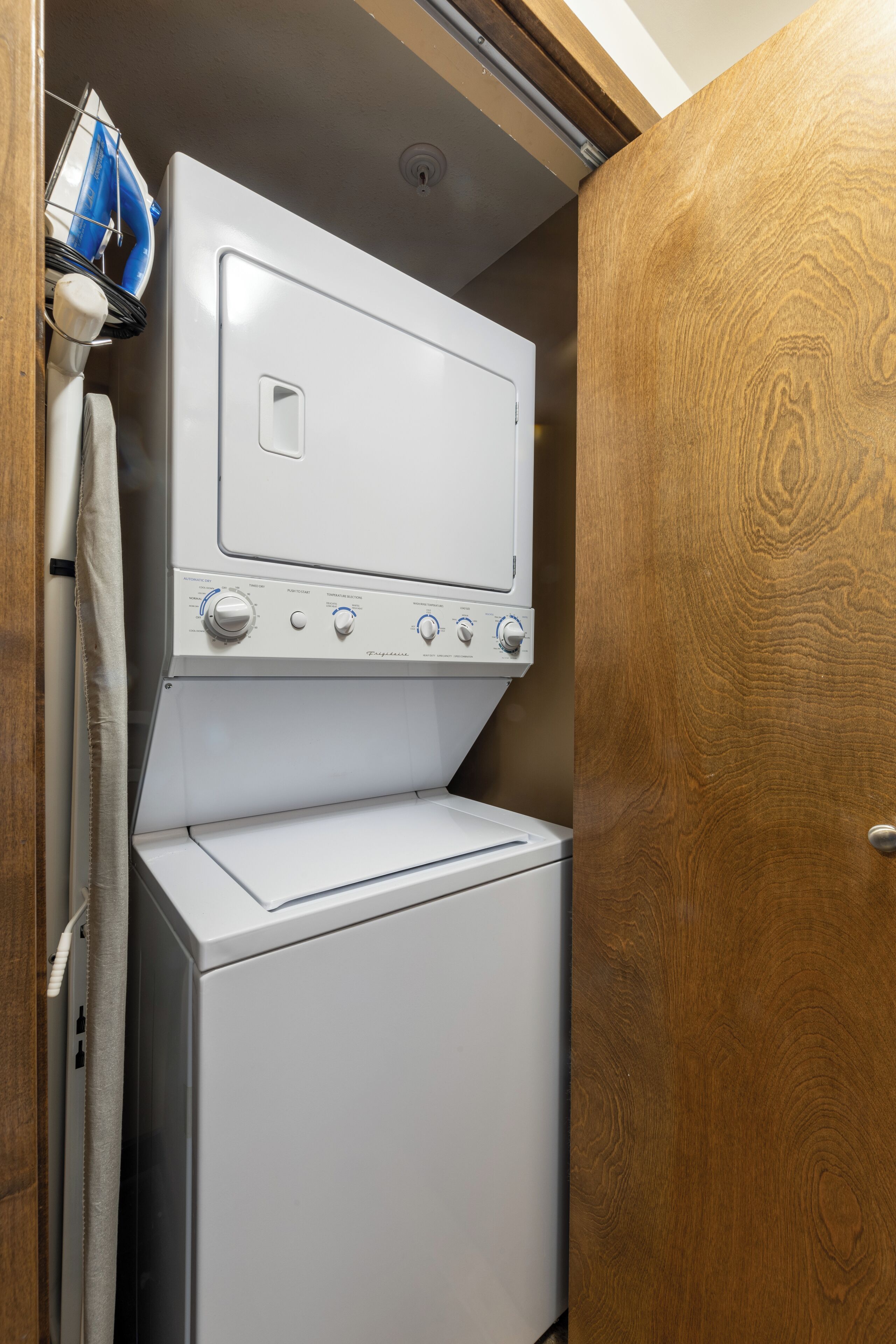A compact laundry area with a front-loading washer and dryer, a wooden door, and a ceiling vent.