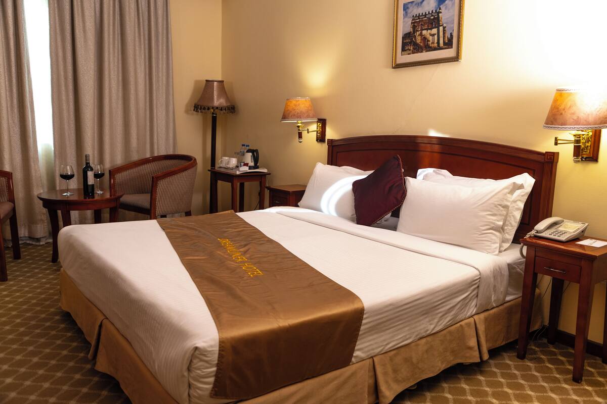 A hotel room with a large bed, a bedside table with a lamp, a chair, and a table with wine glasses and a bottle.