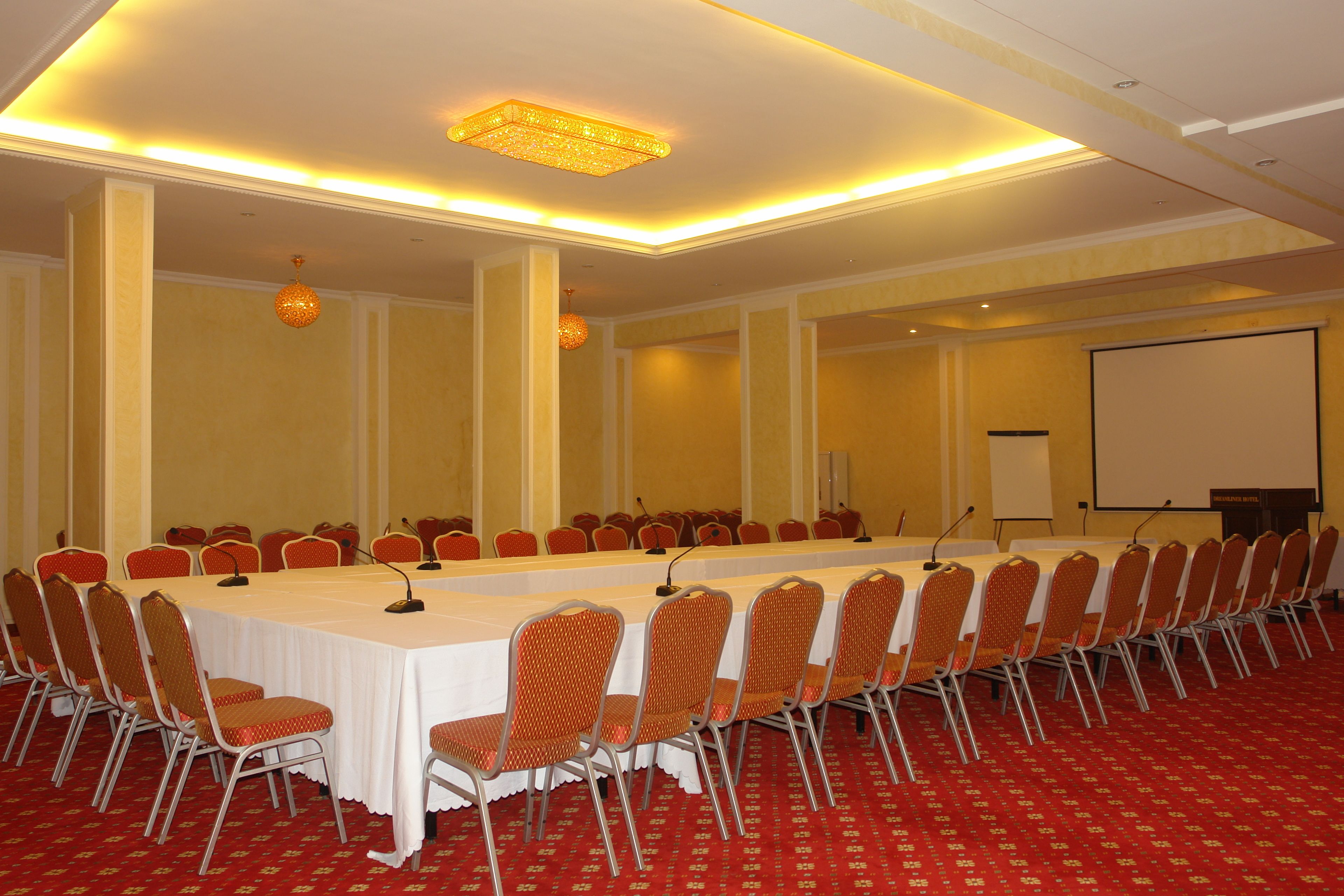 A conference room with a long table, rows of chairs, a projector screen, and a carpeted floor.