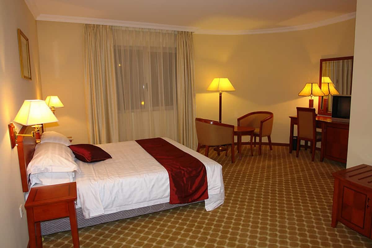 A hotel room with a bed, two bedside tables, a desk with a chair, a television, and a window with curtains.