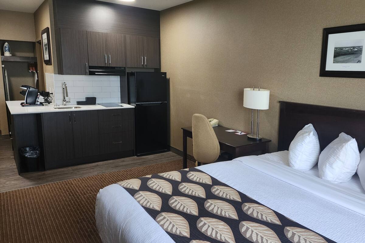A hotel room with a bed, a desk, a chair, a kitchenette, and a framed photo on the wall.