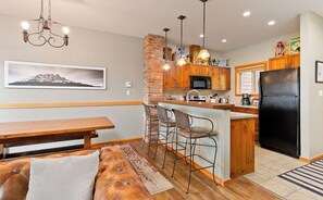 A modern kitchen with a brick accent wall, wooden cabinets, a black refrigerator, and a dining area with a bench and cushioned seating.