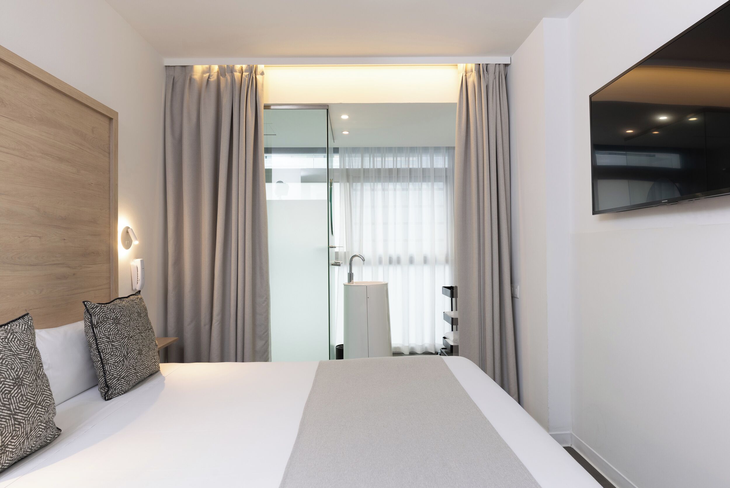 Ein modernes Hotelzimmer mit einem großen Bett, einem an der Wand montierten Fernseher und einem durch eine Glastür sichtbaren Badezimmer.