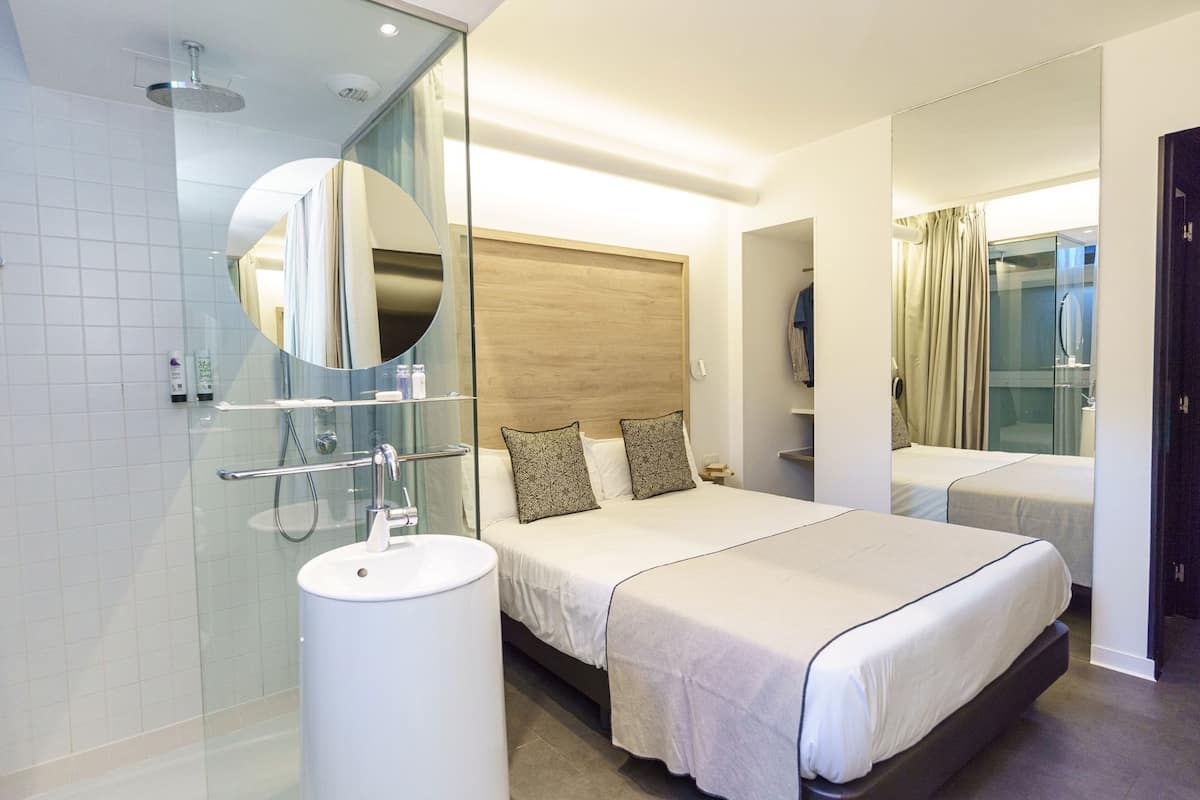 A modern hotel room with a large bed, a separate bathroom, a round mirror, and a glass shower enclosure.