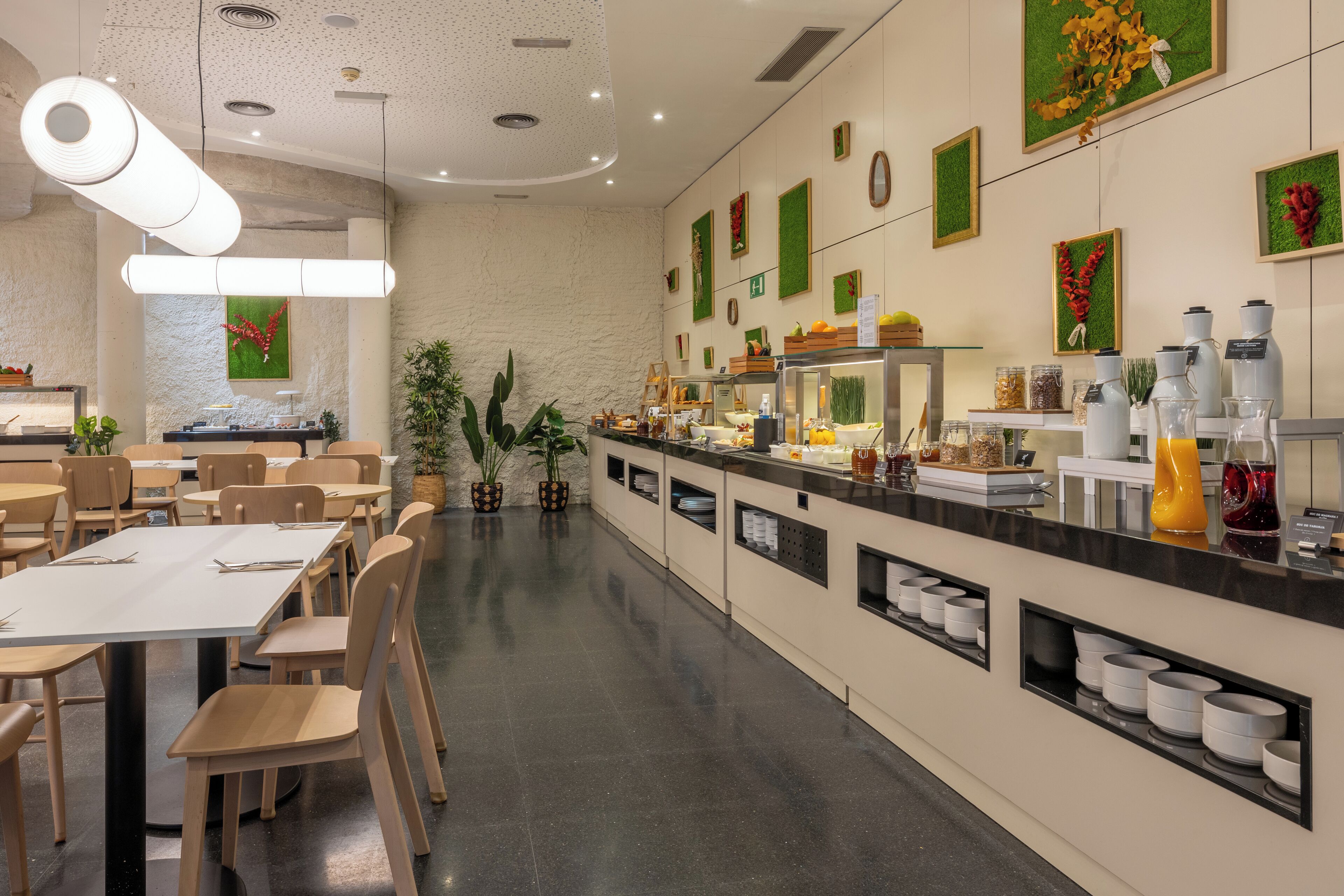 Um restaurante espaçoso com várias mesas e cadeiras, um balcão longo com diversos alimentos e acompanhamentos, e obras de arte decorativas nas paredes.