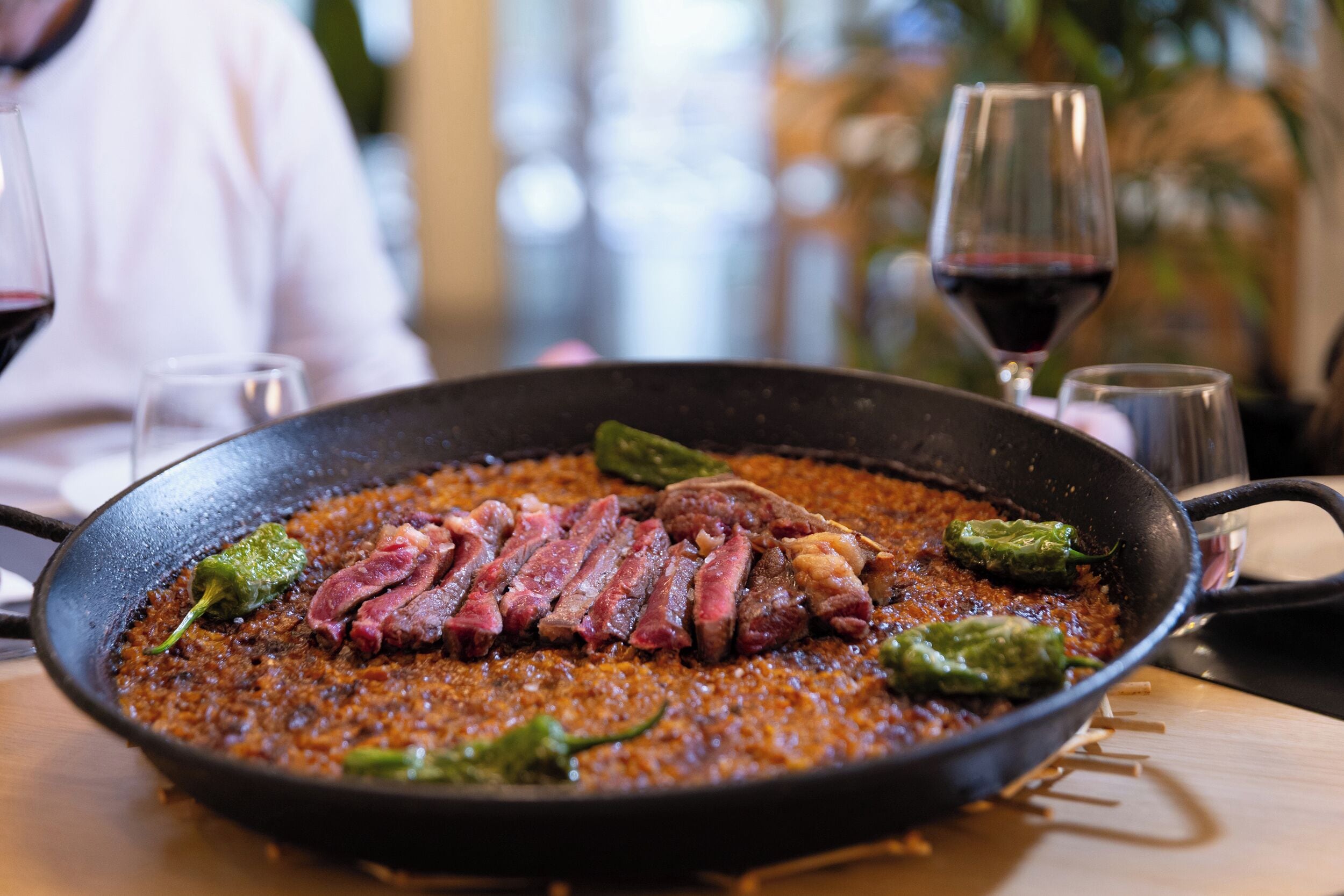 A pan with sliced meat and peppers on a table, with a glass of red wine and a person in the background.