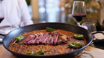 Una sartén con carne y pimientos en una mesa, junto a una copa de vino tinto, y una persona al fondo.