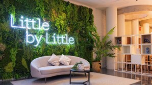 Un interior moderno con un letrero de neón que dice 'Little by Little', un sofá blanco, una mesita con una planta y una estantería blanca.