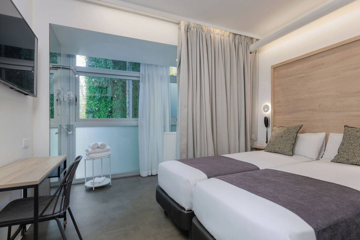 A hotel room with two beds, a desk, a chair, a TV, and a window with curtains.