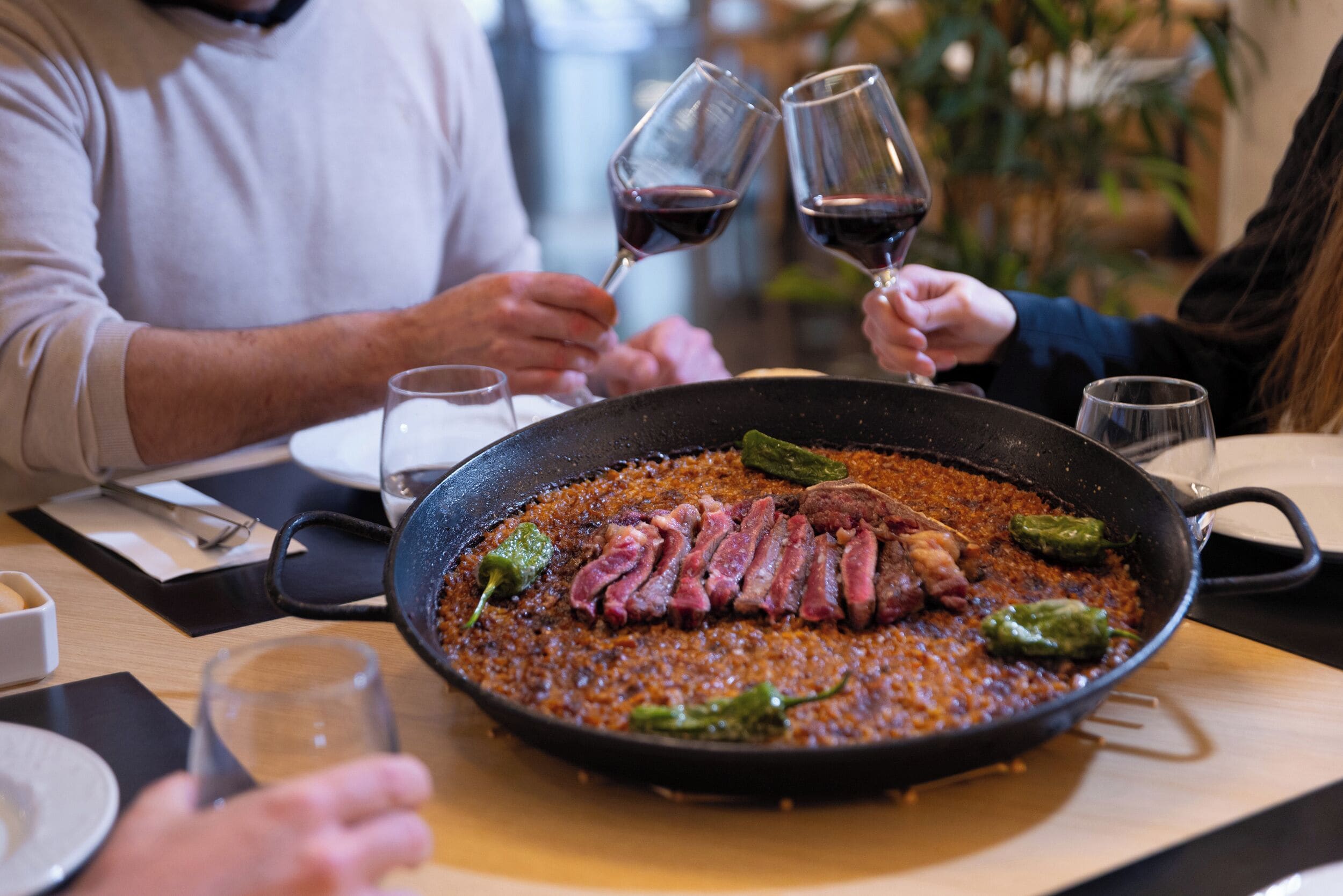 Una mesa de comedor con una bandeja de comida, copas de vino y personas cenando.