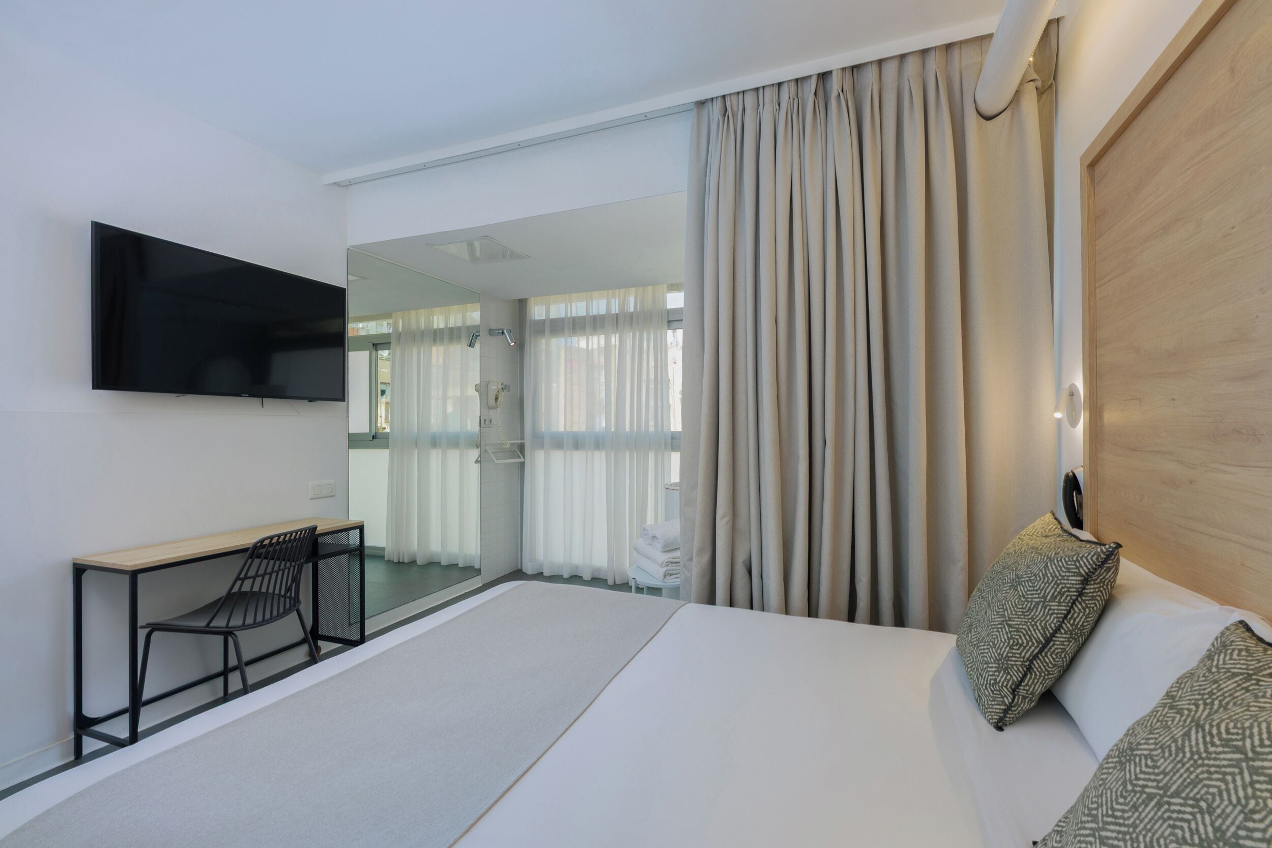 A modern hotel room with a large bed, a desk with a chair, a flat-screen TV mounted on the wall, and a large mirror.