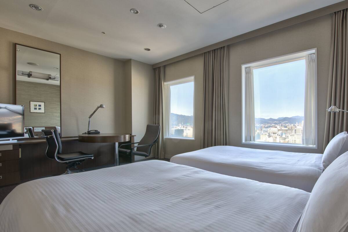 Habitación de hotel con dos camas, un escritorio con silla, una lámpara de escritorio, un espejo y ventanas amplias con vista a la ciudad.