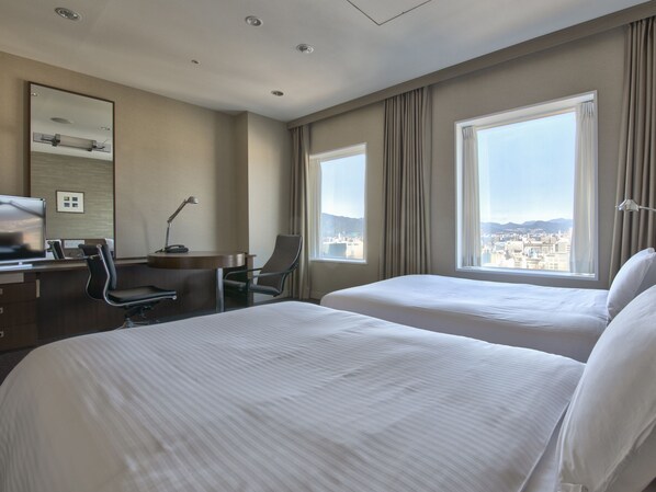 A hotel room with two beds, a desk with a chair, a desk lamp, a mirror, and large windows with a city view.