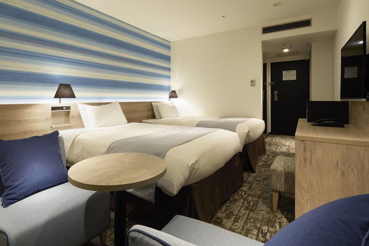 Habitación de hotel con dos camas, una mesita redonda pequeña, un sofá, un televisor y un cuadro abstracto grande en la pared.