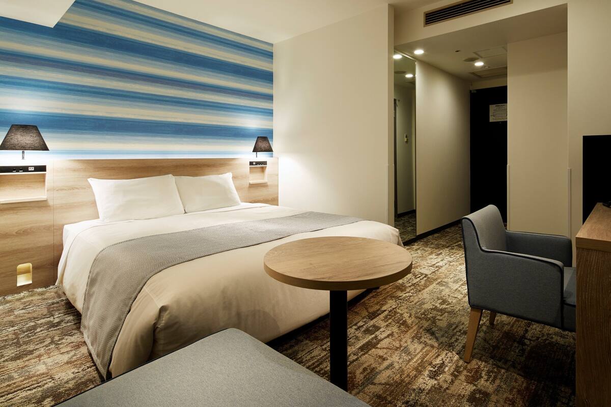 Habitación de hotel con cama, una mesa redonda, una silla, un escritorio, una lámpara y un cuadro en la pared.