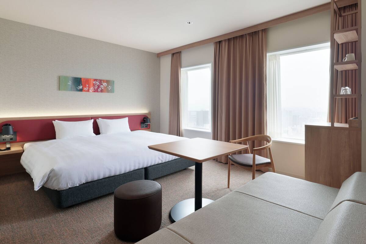 Una habitación de hotel con cama, escritorio, silla, pequeño reposapiés, estantería y ventana con cortinas.