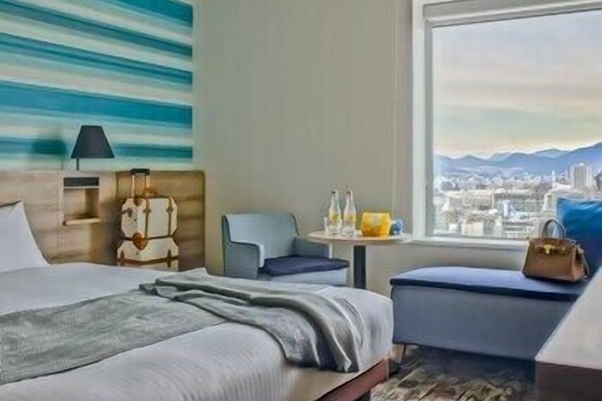 Habitación de hotel con una cama, una silla, una mesita con botellas y vistas a un paisaje urbano a través de un ventanal grande.