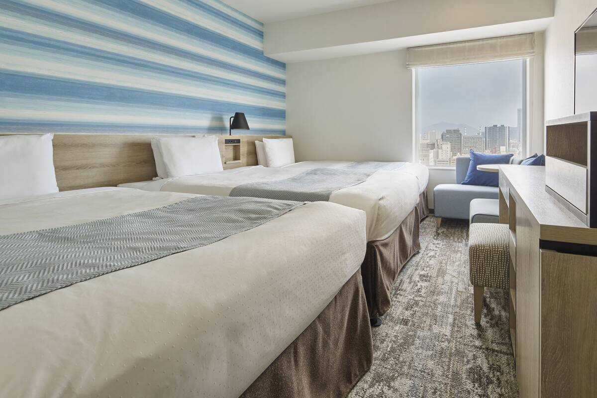 Habitación de hotel con dos camas, un escritorio, una silla y un amplio ventanal con vista a la ciudad.