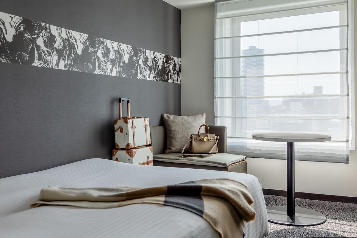 Una habitación de hotel con una cama, una maleta, una mesita y una ventana con persianas.