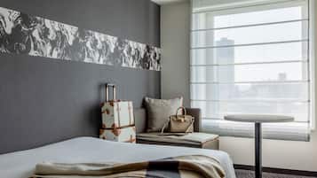 A hotel room with a bed, a suitcase, a small table, and a window with blinds.