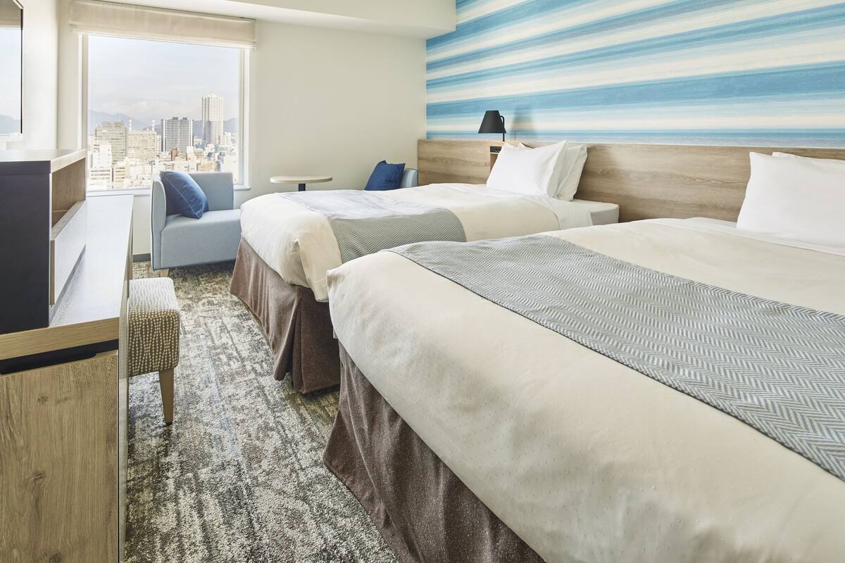 Habitación de hotel con dos camas, un escritorio, una silla y un amplio ventanal con vistas a la ciudad.