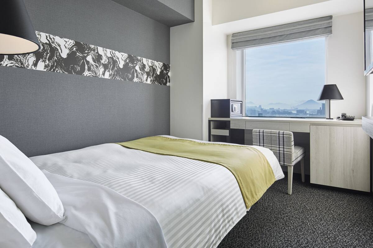 Habitación de hotel con cama, escritorio, silla y ventana que ofrece vistas a la ciudad, con un paisaje urbano visible a través de ella.
