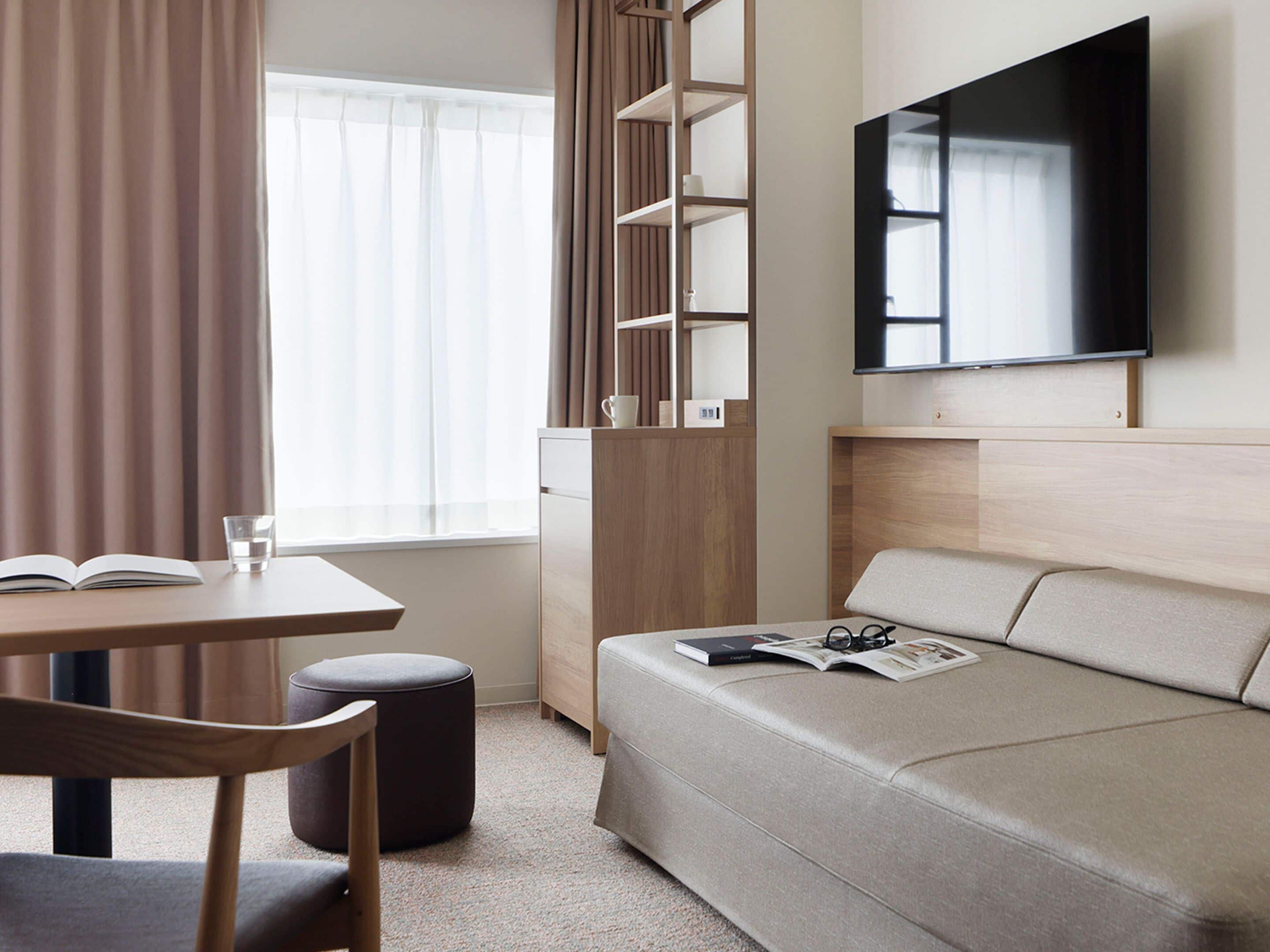 A modern room with a sofa, a table with a book and glass, a TV on the wall, and a window with curtains.